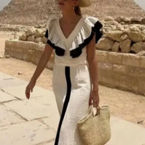 Beige långklänning med volang och svart detalj - Snygg beige långklänning med djup v-ringning och bred volangkant i svart längs axlarna. Klänningen har en svart vertikal rand framtill och är tillverkad i ett lätt linneliknande material. Perfekt för varma dagar och ger en chic vibe.