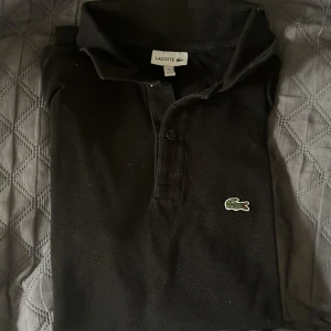 LACOSTE  PIKÉTRÖJA - SVART - STORLEK S/176 CM - Klassisk svart pikétröja från Lacoste med den ikoniska gröna krokodilloggan på bröstet. Tröjan har krage, knäppning med två knappar och är tillverkad i bomull. Perfekt för en clean och stilren look. Äkta vara köpt från Kidsbrandstore.