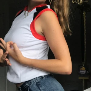 Vit och röd ärmlös linnetopp - Sportig ärmlös linnetopp i vitt med röda och svarta detaljer vid axlar och halsringning. Snygg passform och perfekt till jeans eller shorts. 