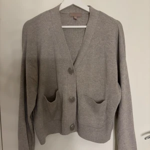 Stickad kofta - Mysig grå kofta med v-ringning och större knappar framtill. Modellen är loose fit med ribbade muddar och två praktiska fickor framtill. Perfekt över en klänning till sommaren, passar xs-m