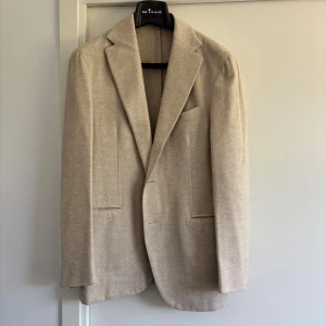 Beige kashmirkavaj från Etro, stl 48 - Exklusiv beige kavaj från Etro i 100% kashmir. Klassisk modell med spetsiga slag, tre bruna knappar vid ärmslut och diskret fiskbensmönster. Fodret har snygga ränder i gult och vitt. Perfekt för dig som gillar stilrena och lyxiga plagg. Nypris runt 22,500kr