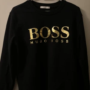 Svart sweatshirt från Hugo Boss - Svart sweatshirt från Hugo Boss med stort guldigt BOSS-tryck på bröstet. Tröjan har rund halsringning och långa ärmar. Perfekt för dig som vill ha en stilren och trendig look med en touch av lyx.