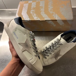 Golden Goose vita sneakers med stjärna - Snygga vita sneakers från Golden Goose med grå mockadetaljer och ikonisk stjärna på sidan. Skorna har grå snörning, mörkblå hälkappa och platt sula. Perfekta för dig som gillar en clean men ändå unik look.