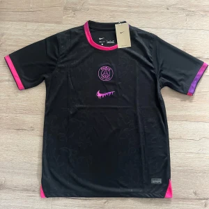 PSG x Nike svart fotbollströja S - Svart PSG fotbollströja från Nike med diskret blommönster i tyget. Rosa och lila detaljer vid krage och ärmslut, samt PSG-logga och Nike Swoosh i rosa framtill. Tillverkad i Dri-FIT-material som andas. Perfekt för dig som vill sticka ut på planen.