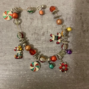 Färgglatt juligt berlockarmband - Charmigt armband med juliga berlocker som pepparkaksgubbe, polkagris, snögubbe och blomma. Pärlor i rött, grönt, gult, lila, orange och silver. Armbandet har glittriga detaljer och blandade former, perfekt för dig som älskar färg och julkänsla.