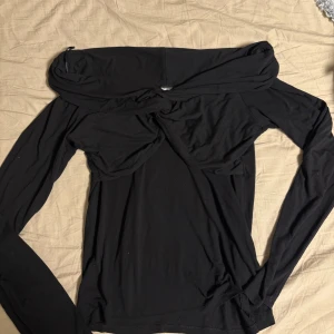 Svart offshoulder topp från Gina Tricot - Säljer en svart offshoulder topp från Gina Tricot i mjuk bomull. Toppen har långa ärmar och en snygg twistad detalj framtill som ger en cool look. Perfekt för dig som gillar trendiga och stilrena plagg med en unik touch. Är lite liten i storleken så passar också L M 