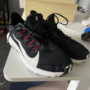 Svarta Nike löparskor med röd detalj - Snygga svarta Nike sneakers med vit sula och stor vit swoosh på sidan. Skorna har röda snörhål och svarta skosnören. sulan har svartvita detaljer. Perfekta för löpning eller träning.