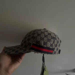 Gucci keps  - Snygg keps från Gucci i beige med klassiskt GG-mönster över hela kepsen. Har en röd och marinblå rand på sidan och justerbar rem baktill. Perfekt accessoar för att lyfta din outfit med lyxig känsla.