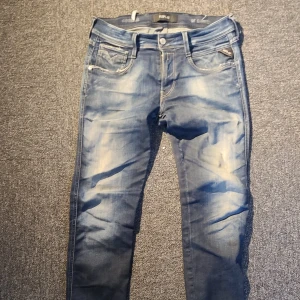 Blå Replay jeans med slitningar - Säljer ett par blå jeans från Replay med snygga slitningar och tvättade detaljer. Klassisk femficksmodell med raka ben och normal passform. Jeansen har tydliga kontrastsömmar och logga på bakfickan. Perfekta för en avslappnad streetstil.