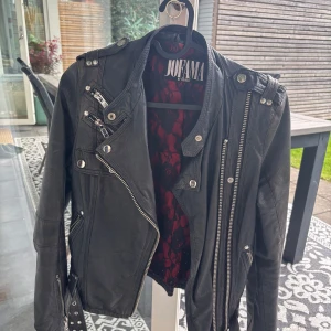 Svart skinnjacka Jofama by Kenza - Säljer en svart skinnjacka från Jofama by Kenza med coola dragkedjor, axelremmar och bälte i midjan. Jackan har lång ärm och rödsvart innerfoder med mönster. Perfekt för dig som vill ha en edgy och tidlös look.