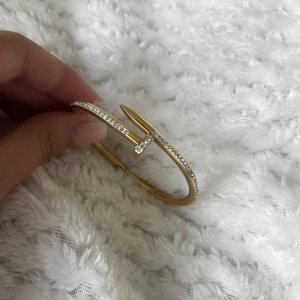 Guldigt spik armband med stenar - Snyggt armband som är format som en böjd spik och dekorerat med glittrande stenar längs sidorna. Armbandet har en unik och modern design som verkligen sticker ut och ger en lyxig känsla. Knappt använd.