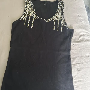 Svart linnetopp med paljetter Amisu - Snygg svart linnetopp från Amisu med massor av silvriga och guldiga paljetter framtill och baktill vid axlarna. Ribbad struktur och ärmlös modell som ger en cool och festlig vibe. Perfekt för dig som vill sticka ut med lite extra bling.