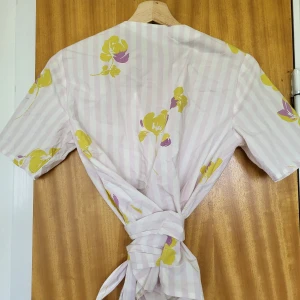 Randig omlottblus Carin Wester 36 - Superfin omlottblus från Carin Wester i ljuslila och vitt med gula och lila blommor. Blusen har kort ärm, v-ringning och långa knytband i midjan. Bra skick och använd bara ett fåtal gånger. 