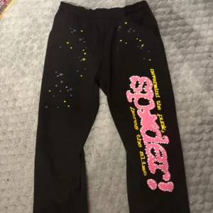 Svarta Sp5der mjukisbyxor med tryck - Svarta mjukisbyxor från Sp5der med elastisk midja och snörning. Stora rosa och vita Sp5der-tryck längs ena benet och små stjärnor i gult, blått och lila. Mjukt bomullsmaterial, perfekt för chill dagar. Snygg streetwear-vibe och unik design.