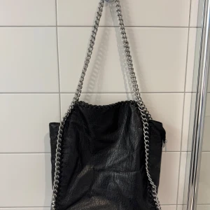 Svart axelväska med kedjedetaljer Tiamo - Cool svart axelväska från Tiamo i skinnimitation med grova silverfärgade kedjor som axelrem och snygg detaljsöm runt kanterna. Väskan har dragkedja både på utsidan och insidan, perfekt för att hålla koll på dina grejer. Köpt på scorett för 599kr.