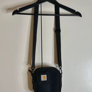 Svart axelväska från Carhartt - Säljer en svart axelväska från Carhartt med justerbar rem och klassisk logga framtill. Väskan har dragkedja och flera fack, perfekt för att bära det viktigaste. Tillverkad i slitstark polyester och har en stilren, kompakt design.
