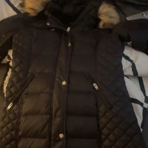 Svart Rockandblue pufferjacka med fuskpäls - Snygg svart pufferjacka från Rockandblue med quiltade detaljer och dragkedjor på fickorna. Jackan har en huva med fuskpäls och Rockandblue-märke på ärmen. Perfekt för kalla dagar och riktigt skön att bära.