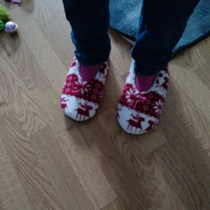 Vita och röda tofflor med julmönster - Mysiga tofflor i vitt och rött med klassiskt julmönster och renar. Insidan är fluffig och mjuk, perfekt för kalla dagar. Utsidan har små rosetter framtill och ett mönster med snöflingor och renar. Sitter skönt på foten och är supervarma.