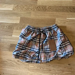 Rutig Burberry shorts med snörning - Snygga shorts från Burberry i klassiskt rutigt mönster med beige, svart, vit och röd. De har elastisk midja med svart snörning och innerbyxa i mesh. Perfekta för sommaren och riktigt ikoniska med Burberry-logga broderad framtill. Passar M och S