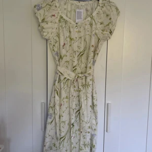 Blommig långklänning från H&M - Superfin långklänning från H&M i ljusbeige med blommigt mönster i grönt, lila och rosa. Klänningen har korta puffärmar, knappar framtill och ett matchande knytbälte i midjan. Perfekt för sommarens alla tillfällen!