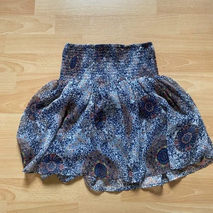 Blommig kortkjol från ONLY - Supersöt kortkjol från ONLY med blått och vitt blommönster, bred smock i midjan och volangig nederkant. Kjolen har ett tunt, luftigt yttertyg och ett mörkblått foder som gör den extra bekväm. Perfekt för dig som gillar boho-vibes och färg!