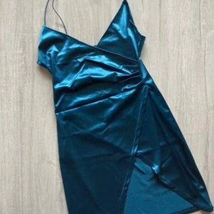 Turkos satin klänning med omlott och smala axelband - Säljer en snygg turkos klänning i satin med smala axelband och omlott-detalj framtill. Klänningen har en figurnära passform och en lätt glansig yta som ger en lyxig känsla. Perfekt för dig som vill sticka ut!