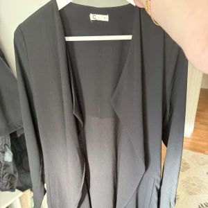 Svart lång kappa från Cubus - Säljer en svart lång kappa från Cubus med öppen front och långa ärmar i storlek S. Kappan har en stilren och minimalistisk design med mjukt fall och bälte i midjan. 