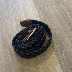 Flätat bälte  - Snyggt flätat bälte i blått, svart och grått med ett stilrent silverfärgat spänne. Bältet har bruna skinnliknande detaljer vid spännet som ger en klassisk touch. Perfekt för att ge din outfit en extra edge!
