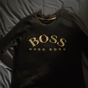 Svart tröja från Hugo Boss - Säljer en svart tröja från Hugo Boss med guldig BOSS-logga framtill i mycket bra skick. Tröjan har rund halsringning och långa ärmar. Perfekt för en stilren och avslappnad look.