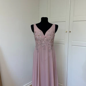 Ljusrosa balklänning med broderade detaljer - Elegant ljusrosa balklänning med v-ringning och vackra broderade blommor med pärlor på överdelen. Klänningen är ärmlös och har en mjukt fallande kjol, perfekt för speciella tillfällen. Dragkedja i ryggen för enkel påklädning.