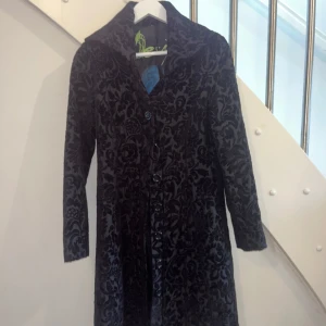 Svart mönstrad kappa från Desigual - Säljer en lång svart kappa från Desigual med vackert blommigt sammetmönster och knappar framtill. Jackan har bred krage och Desigual-logga broderad i blått på ryggen. Fodret är svart med en blå detalj och broderad logga.
