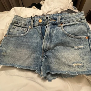 Blå jeansshorts med fransar från bikbok - Säljer ett par blå jeansshorts från bikbok med råa fransade kanter och slitna detaljer framtill. Klassisk femficksmodell med knapp och dragkedja. Perfekta för sommaren!