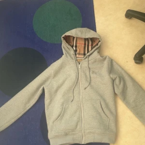 Grå zip-hoodie från Burberry - Säljer en grå hoodie med dragkedja från Burberry. Jackan har klassiskt rutigt foder i huvan, två fickor fram och broderad logga på bröstet. Perfekt för en avslappnad och stilren look.