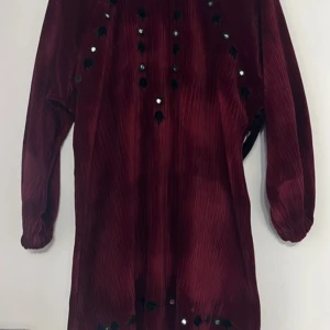 Vinröd långärmad klänning med pärldetaljer - Vinröd klänning med lång ärm och pärldetaljer framtill. Klänningen har en rak passform och är tillverkad i ett mjukt, sammetsliknande material. Perfekt för dig som vill sticka ut med en elegant och unik look.