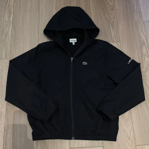 Lacoste blouson - En Lacoste jacka av tunt och lätt material. Vit text på ärmen där det står ”Lacoste”. Perfekt för sommarkvällar.