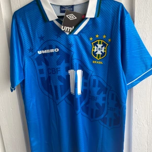 Blå Brasilien fotbollströja Umbro Romario 11 - Säljer en blå Brasilien fotbollströja från Umbro med nummer 11 och Romario på ryggen. Tröjan har korta ärmar, vita detaljer och CBF-logga på bröstet. Perfekt för dig som älskar klassiska landslagströjor.