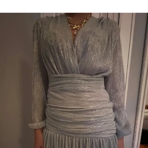 Glittrig silverklänning med omlott och lång ärm - Säljer en glittrig silverfärgad kortklänning med omlott framtill och markerad midja. Klänningen har långa ärmar, v-ringning och plisserad kjol. Perfekt för dig som vill sticka ut med en elegant och trendig look.