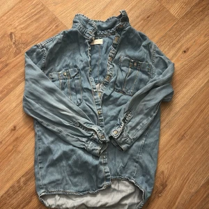 Blå jeansskjorta från Zara - Säljer en klassisk blå jeansskjorta från Zara med två bröstfickor och knappar framtill. Skjortan har lång ärm och avslappnad passform, perfekt till jeans eller kjol. Snygga sömmar och detaljer i kontrastfärg.