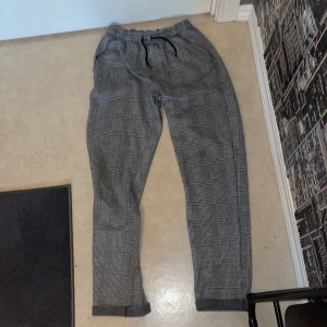 Grå-rutiga byxor från Kappahl - Snygga grå-rutiga byxor från Kappahl med elastisk midja. Byxorna har ett klassiskt rutmönster och raka ben, perfekta för en stilren look. Materialet är en blandning av polyester, viskos och elastan för extra komfort.