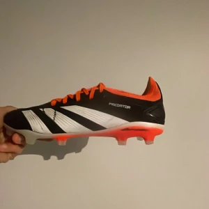 Adidas Predator fotbollssko - Säljer en Adidas Predator fotbollssko med svart ovandel, vita detaljer och orange snörning samt sula. Skon har texten 'PREDATOR' och 'HYBRIDTOUCH 2.0' på sidorna. Perfekt för dig som vill sticka ut på planen. Knappt använda 
