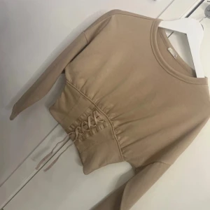 Beige croppad tröja med snörning - Säljer en beige croppad tröja med långa ärmar och snygg snörningsdetalj framtill i midjan. Tröjan har en rund halsringning och är tillverkad i mjukt bomullsmaterial. Perfekt för en trendig och avslappnad look.