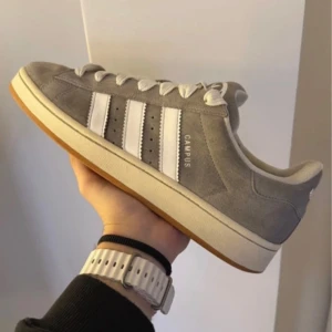 Grå Adidas Campus sneakers - Säljer ett par grå Adidas Campus sneakers med vita ränder och gummisula. Skorna har klassisk låg profil, rund tå och är tillverkade i mocka med vita detaljer och snörning. Perfekta för en stilren vardagslook.