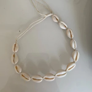 Snäckskalshalsband med justerbart band - Ett handgjort halsband med vita snäckskal trädda på en vit tråd. Halsbandet har justerbar längd och ger en strandig känsla. Perfekt accessoar för sommaren och festivaler.