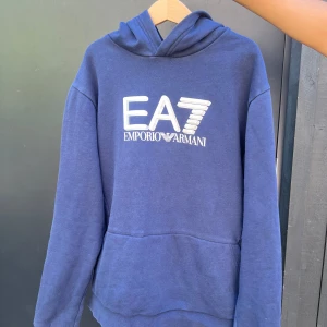 Blå hoodie från Emporio Armani EA7 - Säljer en blå hoodie från Emporio Armani EA7 med stor vit logga på bröstet. Tröjan har huva och en stor magficka framtill. Perfekt för dig som gillar stilrena och bekväma plagg. 