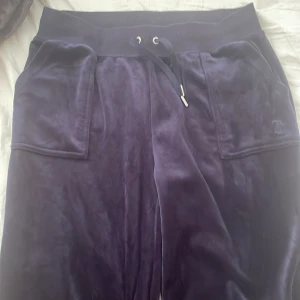 Mörklila velourbyxor från Juicy Couture - Säljer ett par marinblåa velourbyxor från Juicy Couture. Köpte fel storlek det är därför jag säljer dom. Dom har bred resår i midjan och snörning. Byxorna har stora fickor fram och bak med knappar och klassisk Juicy Couture-logga på ena fickan. Perfekta för en avslappnad och trendig look.