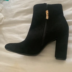 Svarta mocka boots med klack och dragkedja - Säljer ett par svarta boots i mocka med hög klack och spetsig tå. Skorna har en snygg guldfärgad dragkedja på sidan som detalj. Perfekta för att lyfta din outfit med en stilren och trendig look.