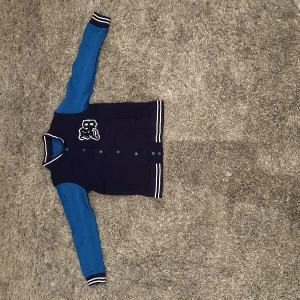 Blå och marinblå bomberjacka med tryck - Snygg bomberjacka i collegestil med marinblå kropp och blå ärmar. Jackan har vita ränder på muddar och krage, knappar framtill och ett vitt tryck på bröstet. Perfekt för en sportig look.