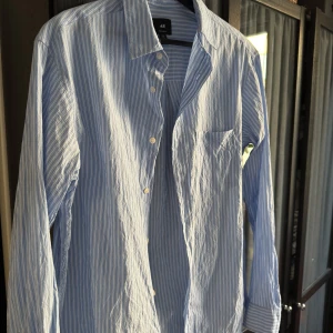 Blåvit randig skjorta från H&M - Klassisk blå och vit randig skjorta från H&M med regular fit. Skjortan har lång ärm, knappar framtill och en bröstficka. Perfekt för en stilren och avslappnad look.