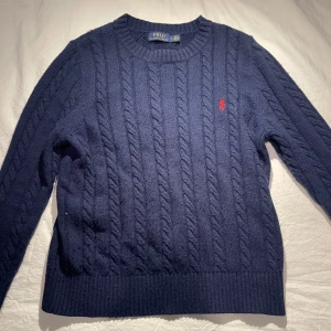 Marinblå kabelstickad tröja från Polo Ralph Lauren - Säljer en klassisk marinblå kabelstickad tröja från Polo Ralph Lauren med rund halsringning och röd broderad logga på bröstet. Tröjan har långa ärmar och ribbade muddar vid ärmslut och nederkant. Perfekt för lager-på-lager-stil. Riktigt fet stickad tröja som man kan ha året runt men skulle säga att den passar bäst under vintern. Nypris ≈ 2400kr. Använd max 20 gånger och i nästan ny skick. Priset går att pruta! Om du har någon fundering så är det bara att skriva till mig och fråga!