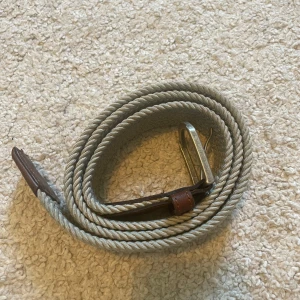 Beige Flätat Bälte - Snyggt beige bälte med flätad design och detaljer i brunt skinn. Bältet har ett klassiskt metallspänne och passar perfekt till jeans eller chinos. Stilren och enkel look som funkar året runt.